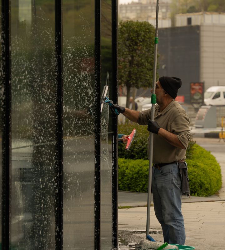 A window cleaner working a water-fed pole up a tall glass frontage, bucket at his feet (stock image)