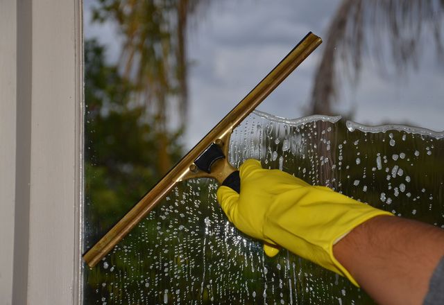 A gloved hand drawing a squeegee across a wet window with trees reflected in the glass